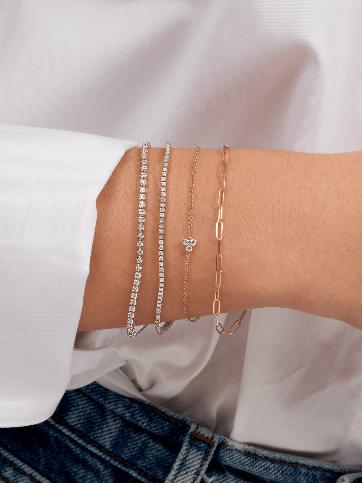 Bezeled diamond tennis chain bracelet in white gold layered with dainty tennis bracelet, gold paperclip bracelet, and diamond trio bracelet with dainty gold chain