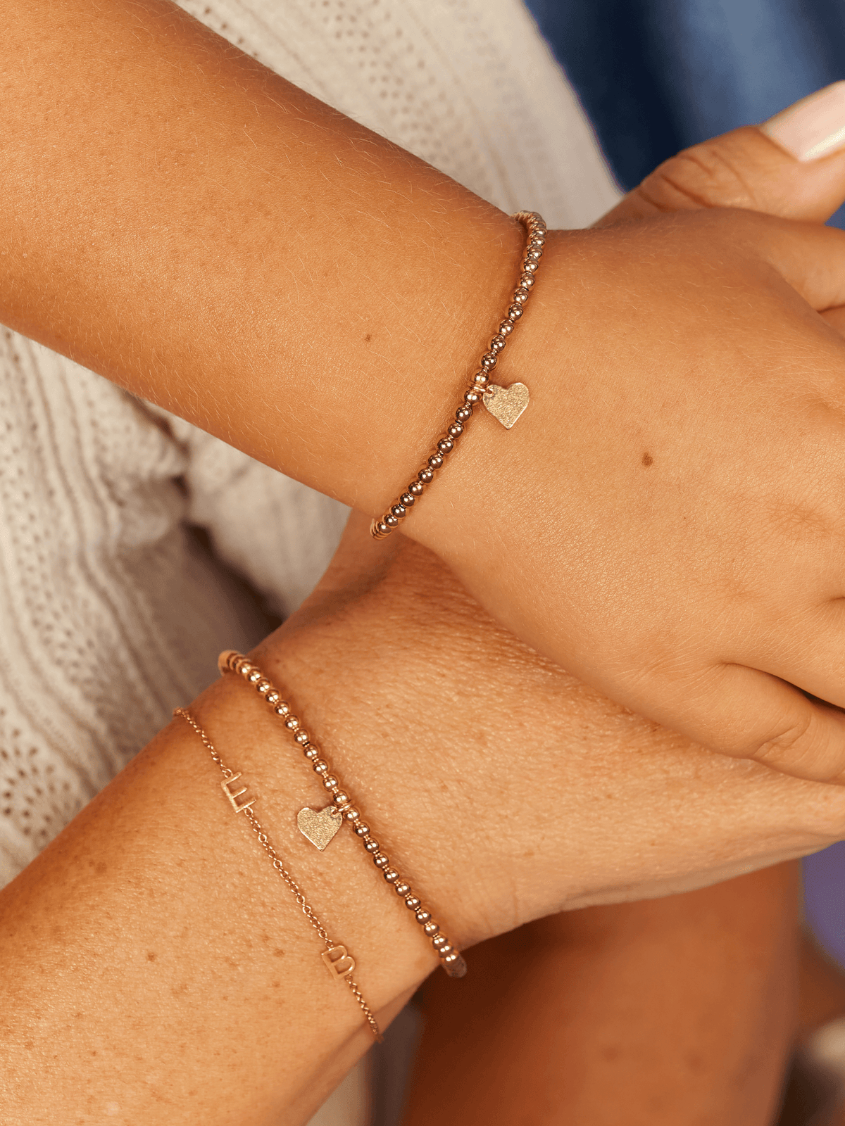 Stretch beaded bracelet with heart charm on top model's wrist and stretch beaded bracelet with heart charm stacked with gold initial bracelet on bottom model's wrist