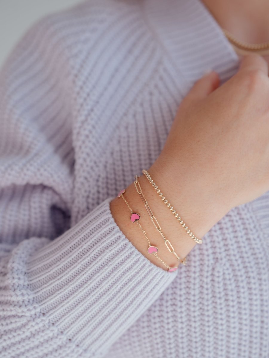 Gold bead stretch bracelet paired with gold paperclip bracelet and pink enamel heart bracelet on model wrist