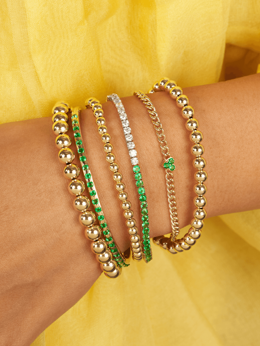Gold beaded bracelets paired with emerald cuff, diamond and emerald tennis bracelet, and emerald heart curb chain bracelet on model wrist