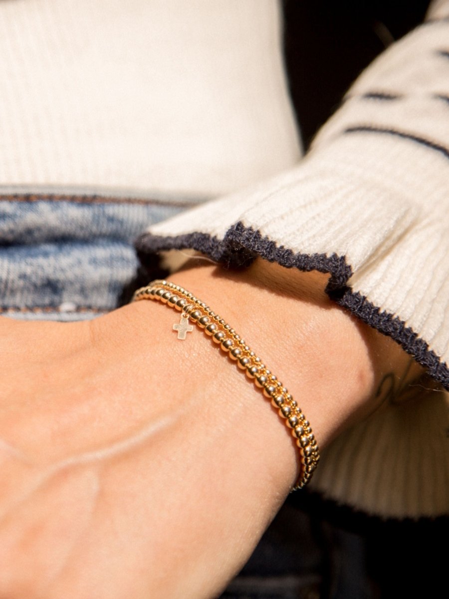 Gold beaded stretch bracelet with cross charm paired with thin gold beaded bracelet on model wrist