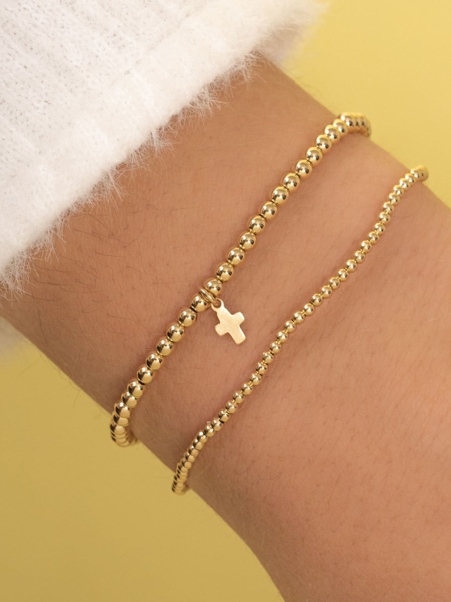 Gold beaded stretch bracelet with cross charm paired with thin gold beaded bracelet on model wrist