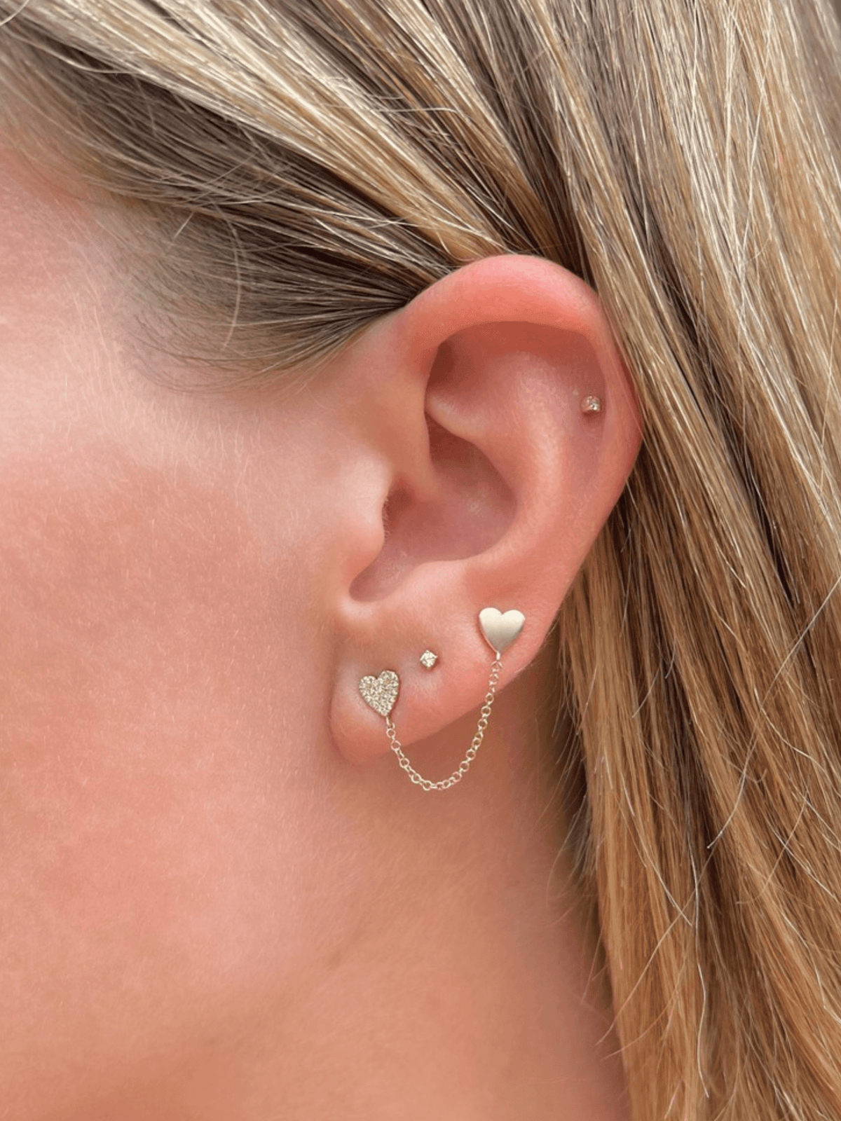 Yellow gold heart stud earring attached to pave heart stud earring linked by chain on model ear with two tiny diamond stud earrings also on ear
