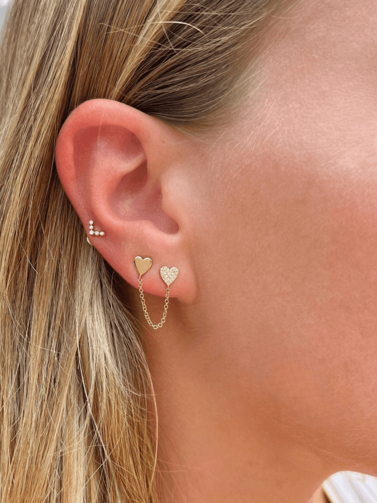 Yellow gold heart stud earring attached to pave heart stud earring linked by chain on model ear with boomerang stud earring 