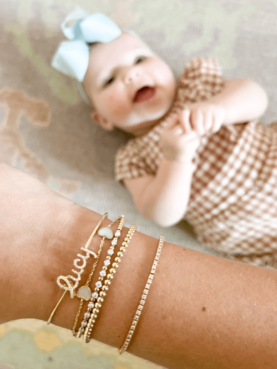 14K gold diamond name bangle in yellow gold layered with gold beaded bracelet, mother of pearl heart charm bracelet, and other gold and diamond bracelets